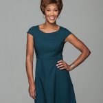 Smiling woman with short curly wig in teal dress, posing confidently.