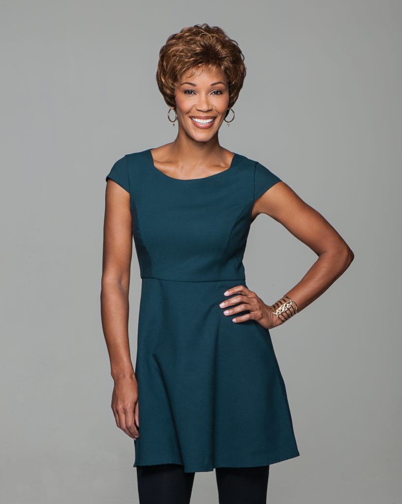 Smiling woman with short curly wig in teal dress, posing confidently.