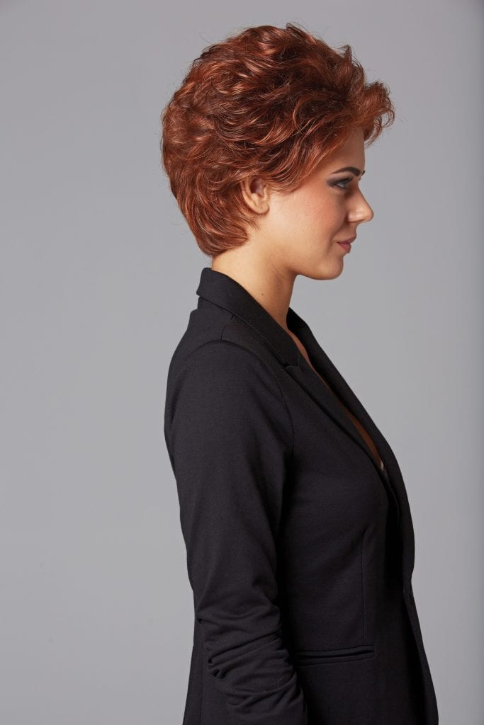 Side view of a woman with short, curly, auburn wig and black blazer.