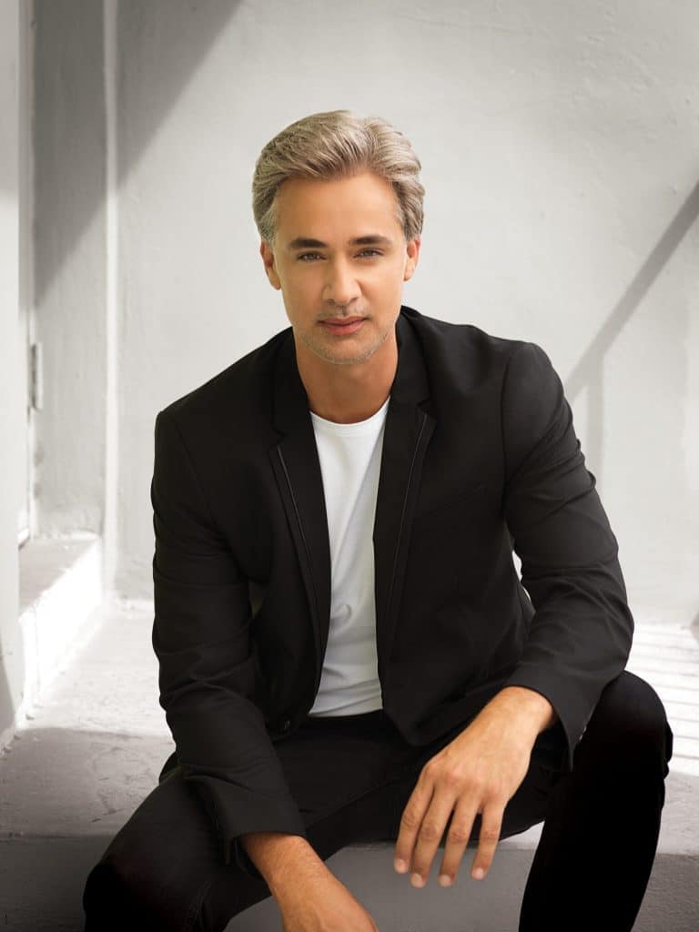 Man with styled gray wig in black blazer and white shirt.
