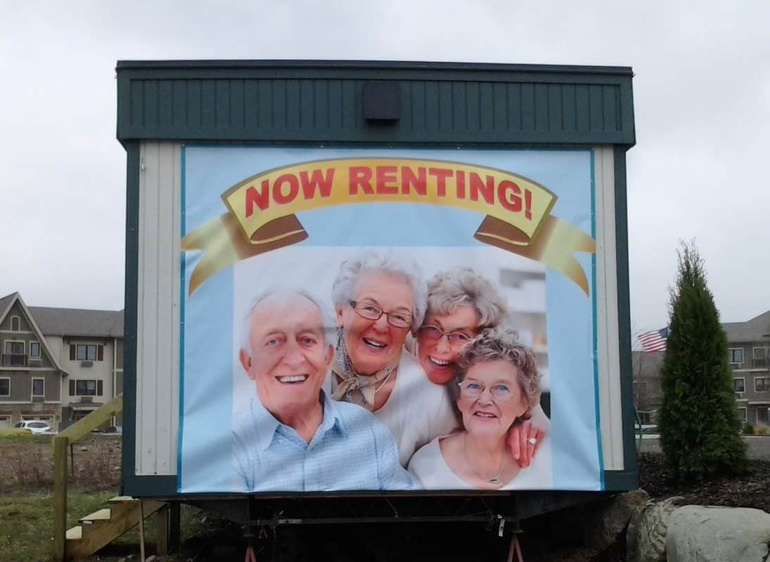 Now Renting sign featuring smiling elderly people in front of a residential area.