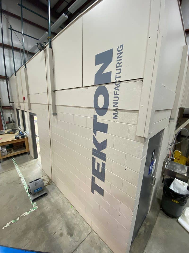 Large industrial manufacturing machine with Tekton logo in factory setting.