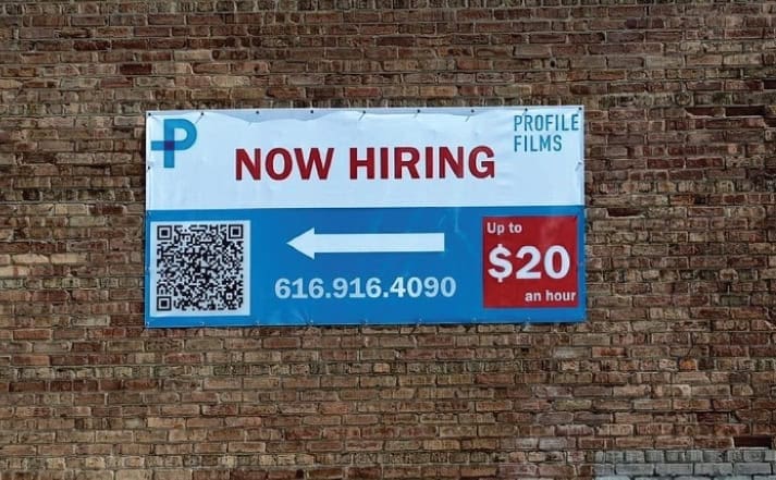 Now hiring sign on brick wall with QR code and hourly wage details.