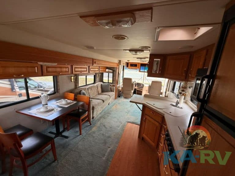 Interior view of an RV showing a cozy living area, kitchen, and dining space with wooden cabinets an.
