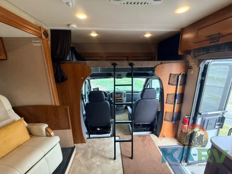 RV interior showing driver and passenger seats with ladder and living area.