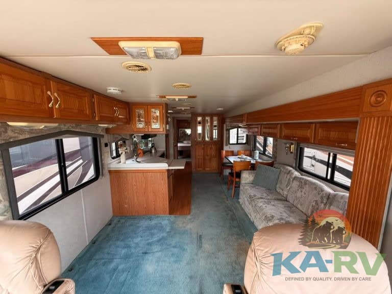 Spacious RV living room with large windows, cozy sofa, and kitchen in the background.