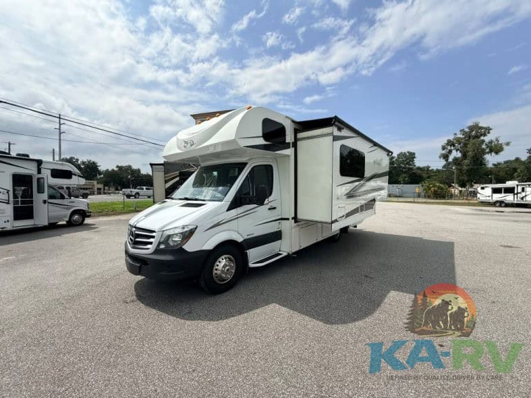 White Class C RV with slide-out parked on lot under blue sky.