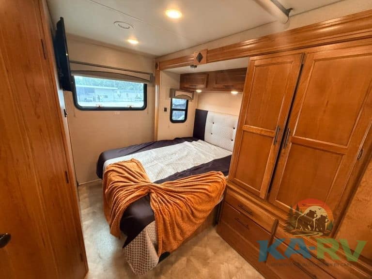 Comfortable RV bedroom with bed, windows, and wooden storage cabinets.