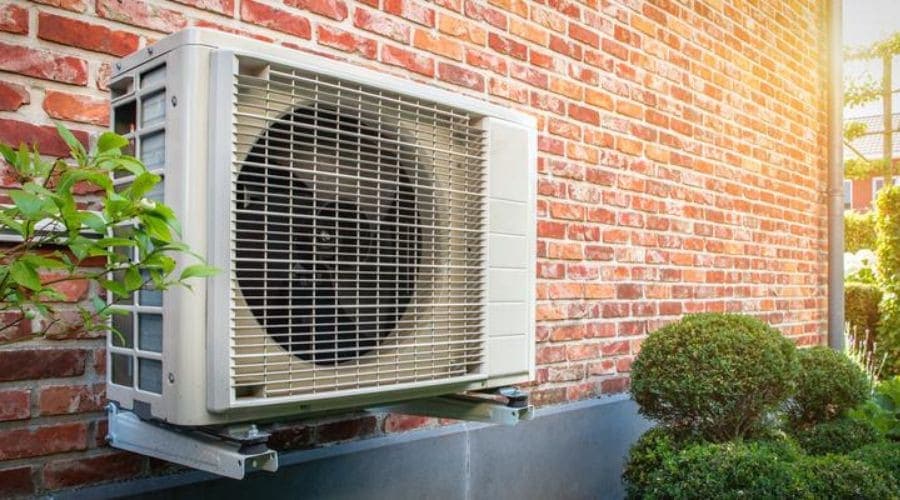 Wall-mounted outdoor air conditioning unit on brick wall with greenery.