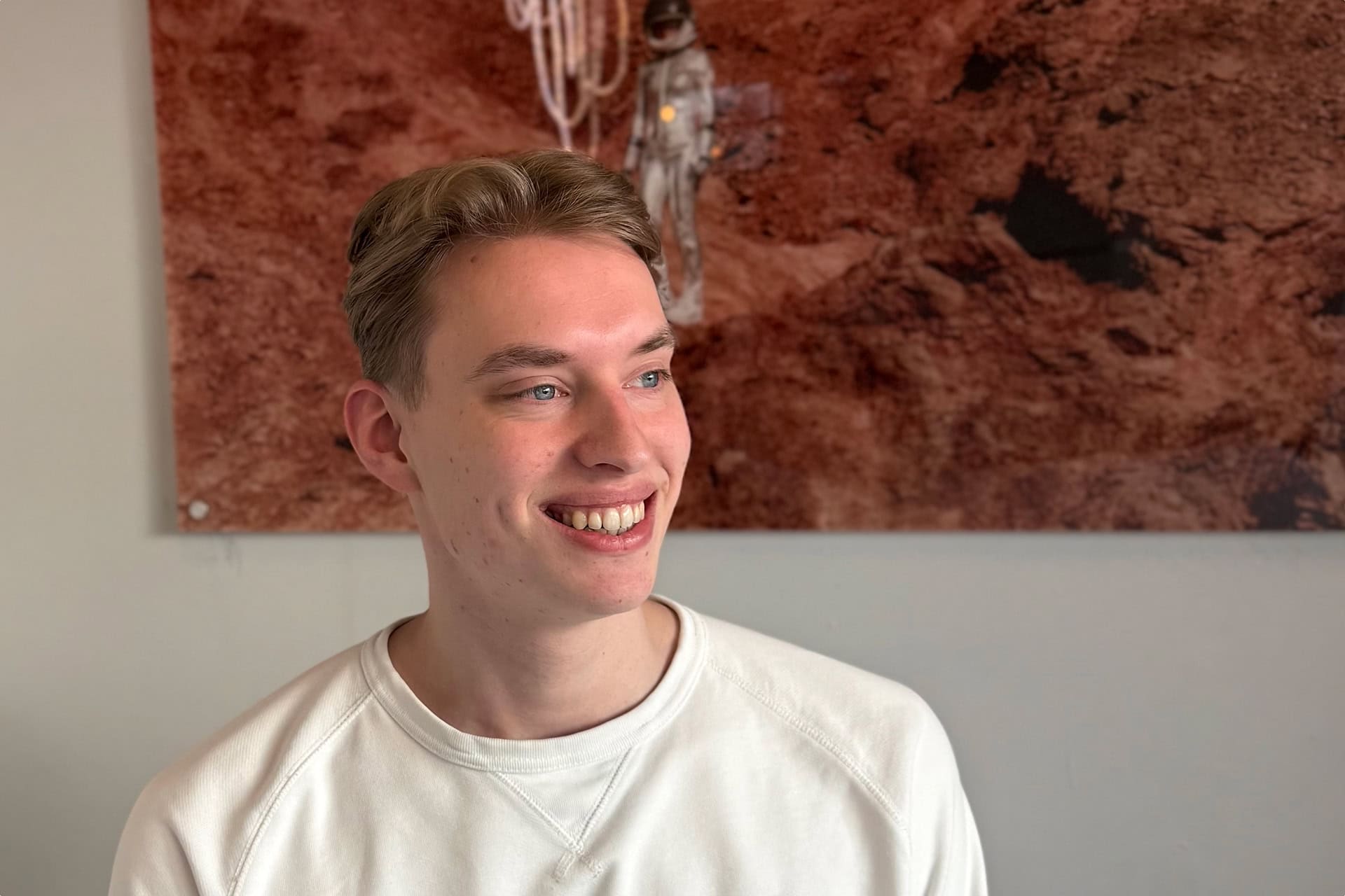 Jonge man in wit sweatshirt glimlacht en kijkt naar rechts met een roodbruine gestructureerde achtergrond.