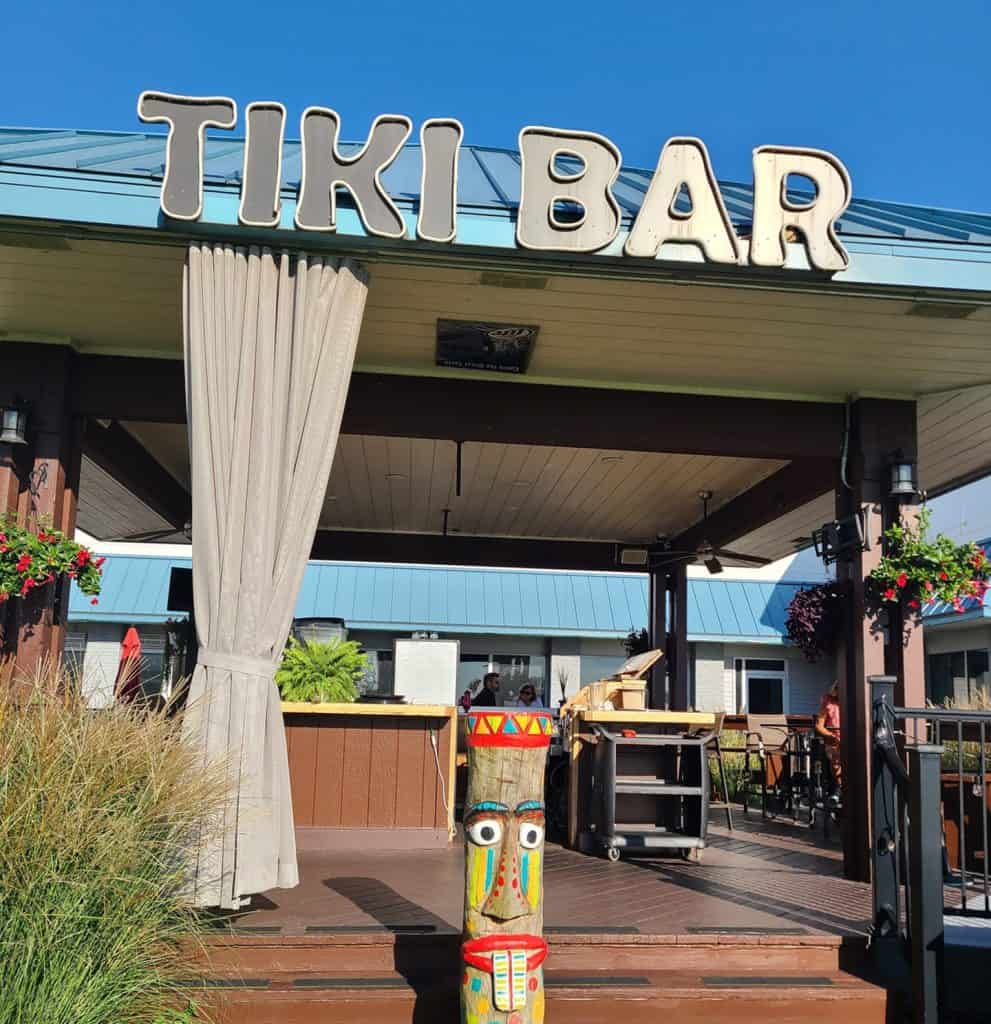 Tiki Bar entrance with colorful tiki statue, outdoor seating, and tropical decor at Mudpenny restaur.