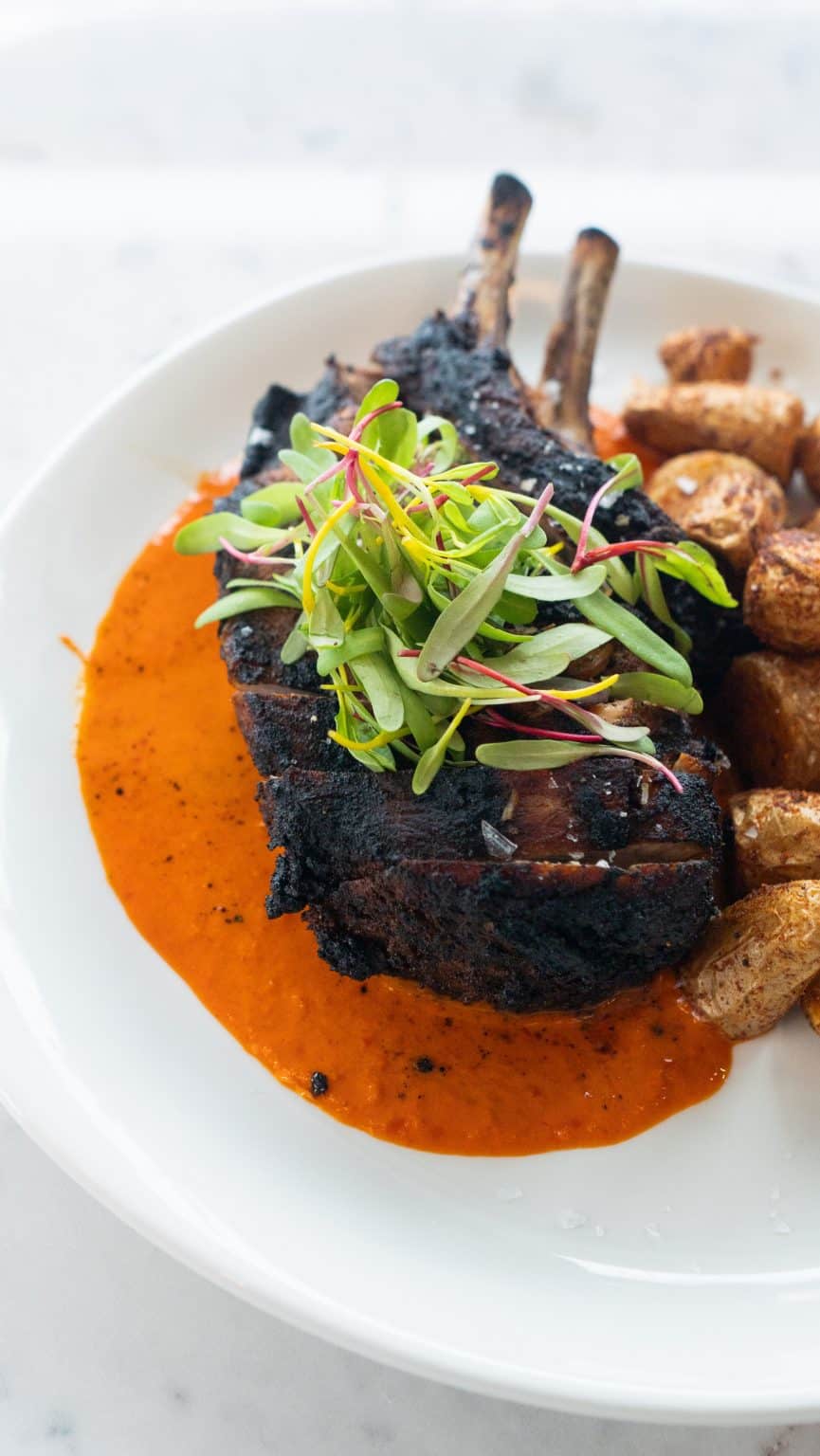 Juicy grilled lamb chops with fresh greens, roasted potatoes, and flavorful sauce on a white plate.