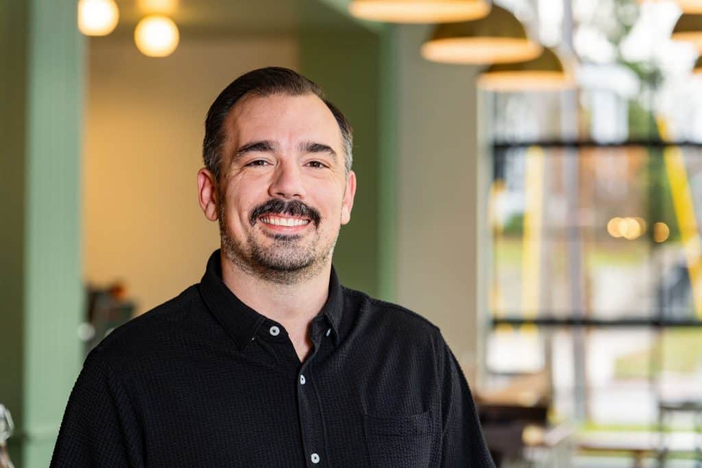 Smiling man in a black shirt at a welcoming cafe, showcasing friendly atmosphere and modern interior.