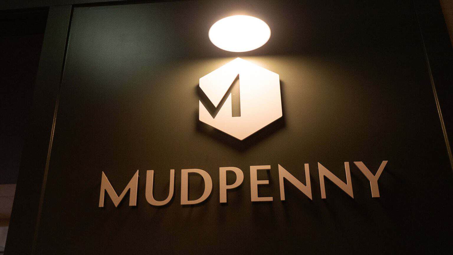 Mudpenny logo illuminated on a dark wall, showcasing modern branding.