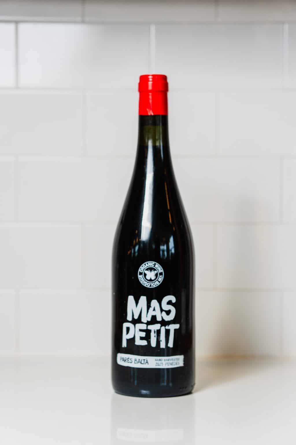 Red wine bottle with Mas Petit label and red cap on white background.