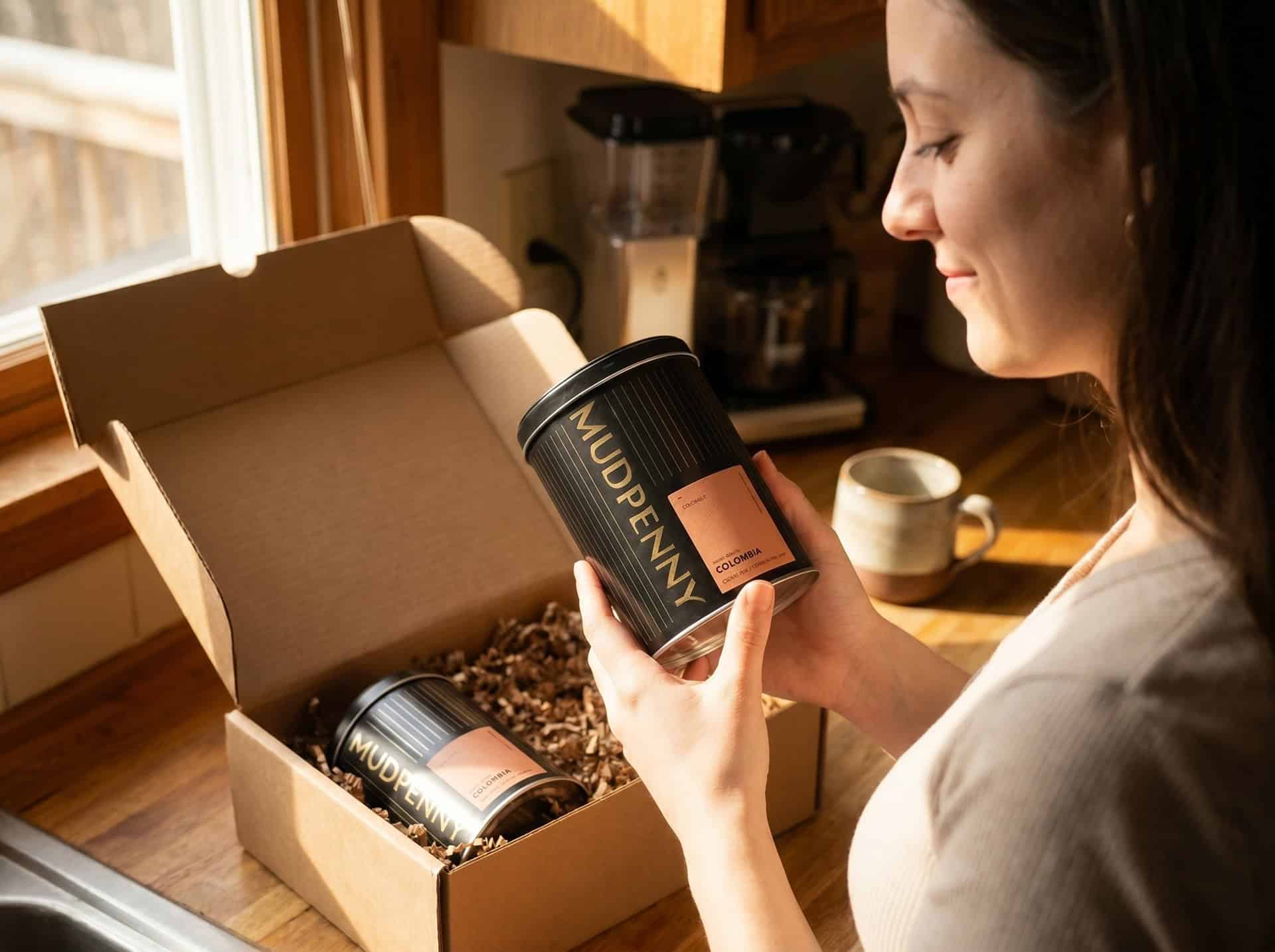 Woman holding Mudpenny coffee container with subscription box on kitchen counter.