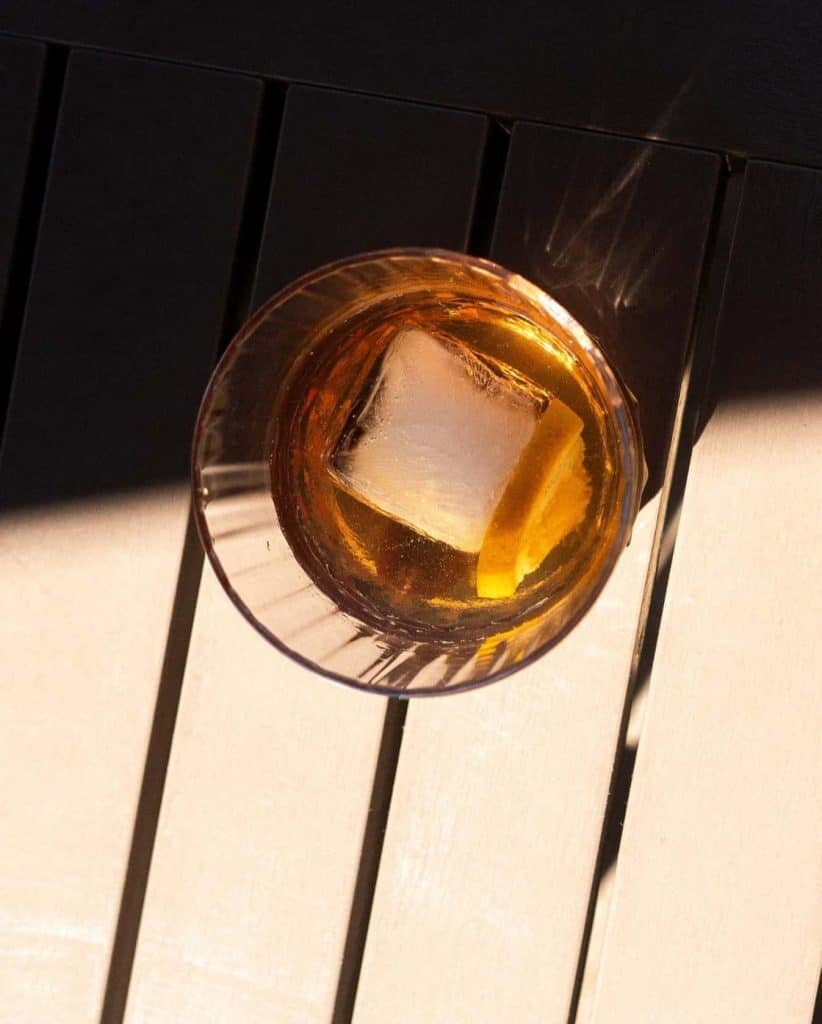 Whiskey glass with ice cube on dark wooden table, top view.