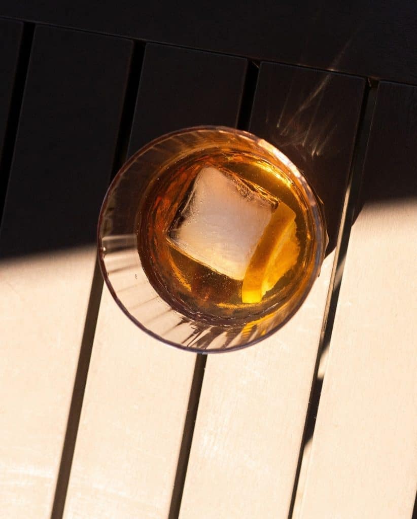 Whiskey glass with ice cube on a dark wooden table, warm lighting highlighting the drink's amber col.