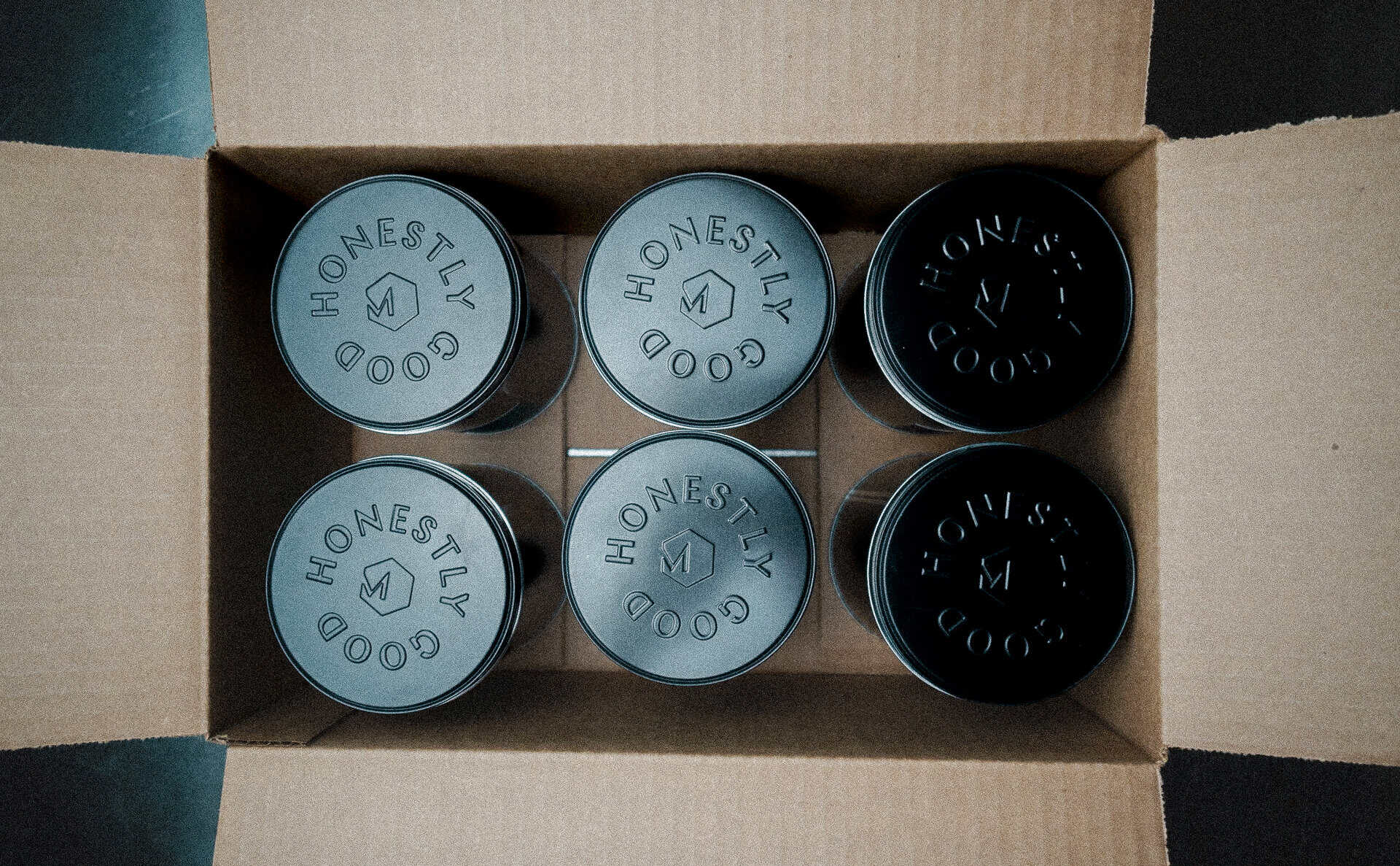Set of six Mudpenny coffee canisters with "Honestly Good" branding, in black and gray, inside a card.