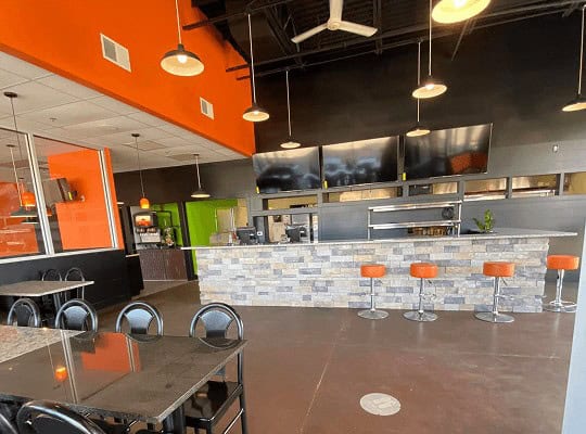  Modern restaurant interior showing the dining area leading to the bar with stainless steel accents, bright orange bar stools, orange wall accents, and white tile bar front.