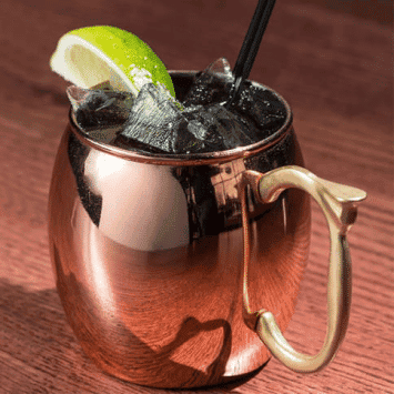 Copper mug cocktail with lime wedge and ice, black straw, on wooden surface.
