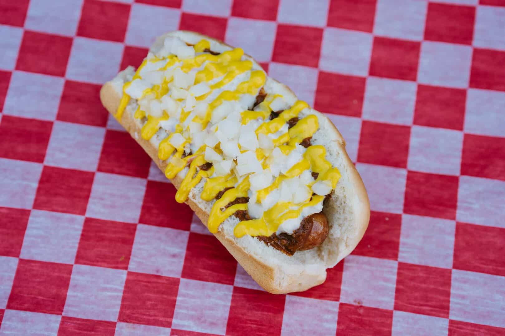 Hot dog with mustard, relish, and chopped onions on a bun on red checkered tablecloth.
