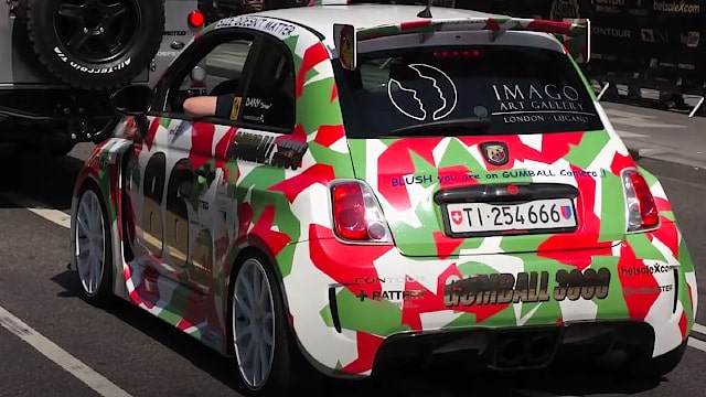 Lille bil med farverig camouflagemønster og mange klistermærker, herunder Gumball 3000 og Imago Art Gallery, set bagfra på en gade.