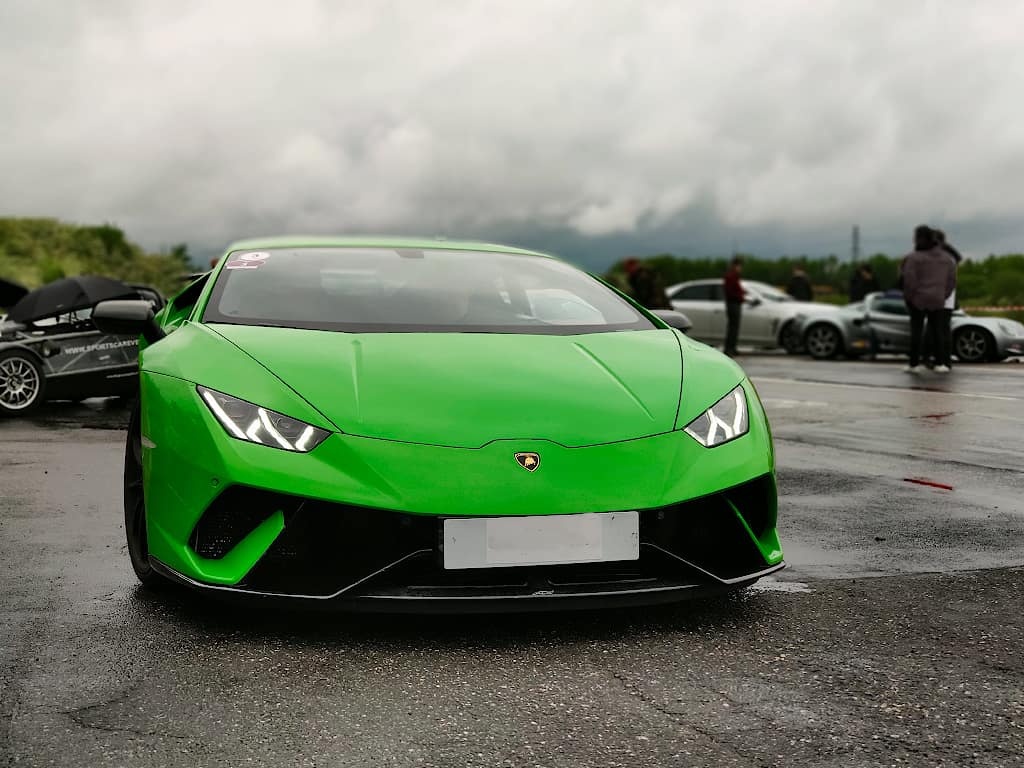 Grøn Lamborghini sportsvogn parkeret på en våd vej med flere biler og mennesker i baggrunden.