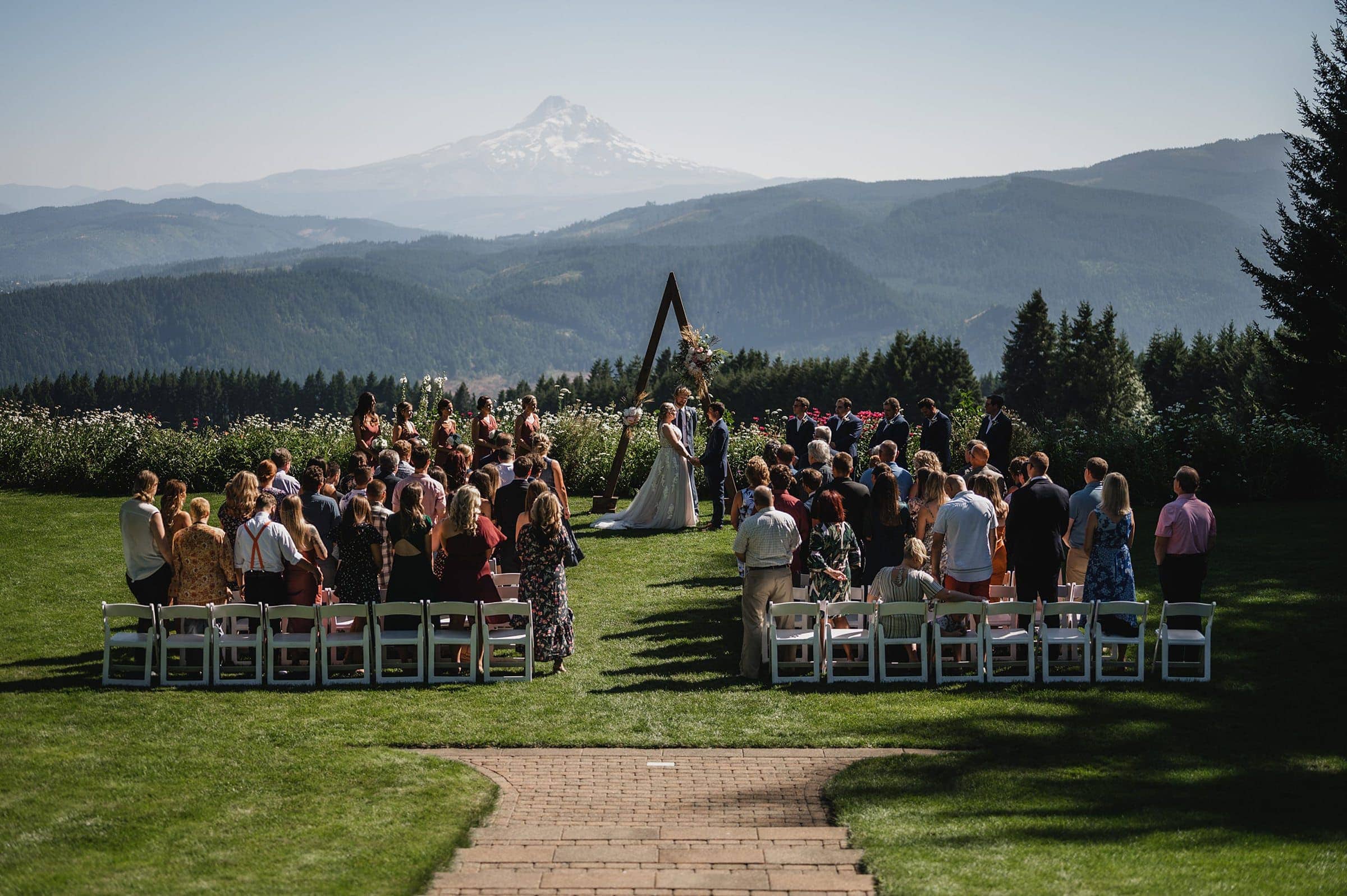 Amidst the stunning mountain and forest backdrop, guests gather on the lawn for a beautiful outdoor wedding ceremony at Gorge Crest Vineyards. The couple stands gracefully under a geometric arch, creating picture-perfect moments captured in exquisite Gorge Crest wedding photos.