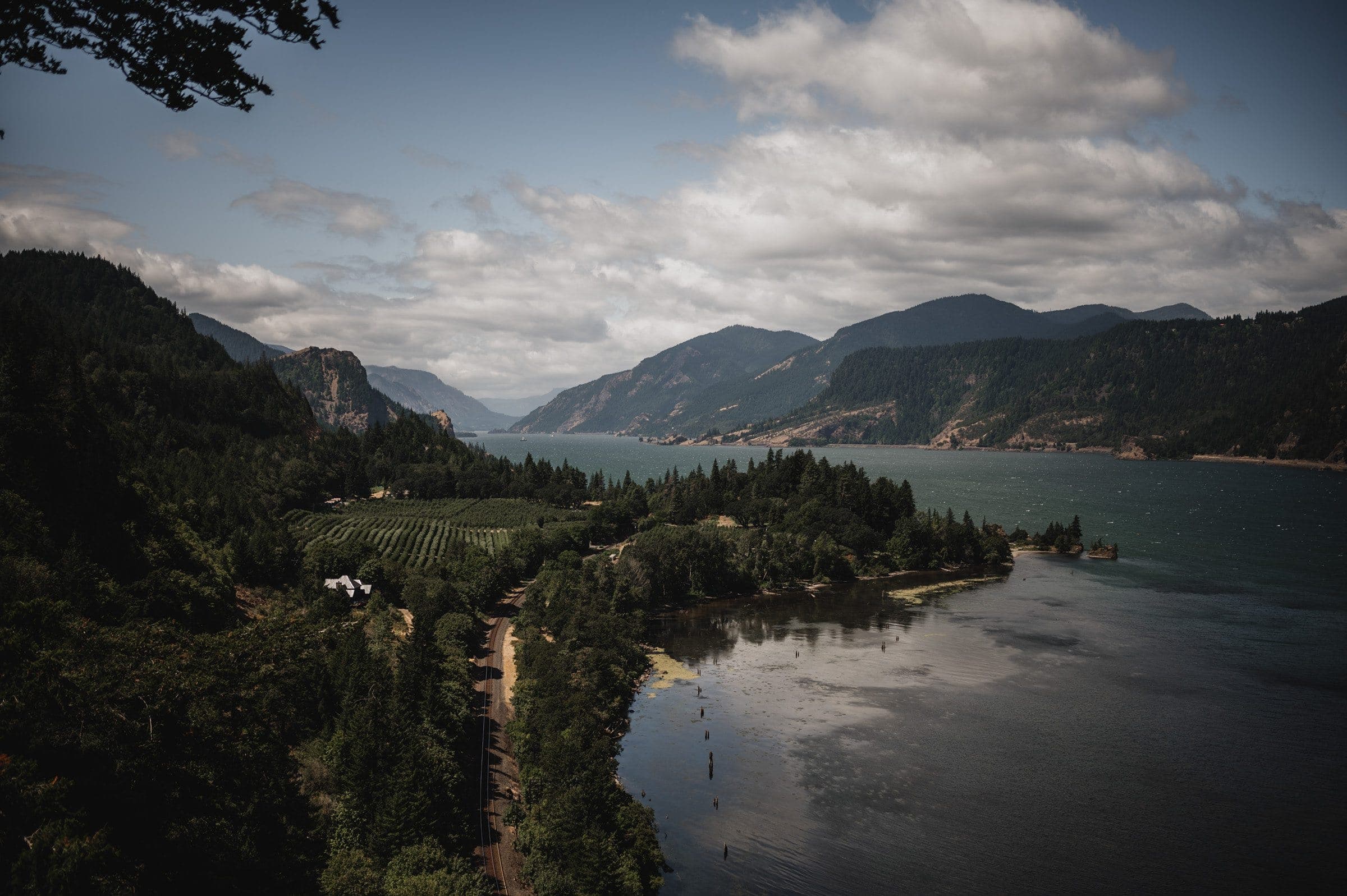 The Columbia River flows through a lush valley with forested hills and majestic mountains under a cloudy blue sky, as view at a wedding photographed at The Griffin House.