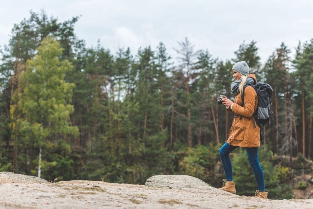 side view of young woman photo camera in hands standing in rock in autumn forest