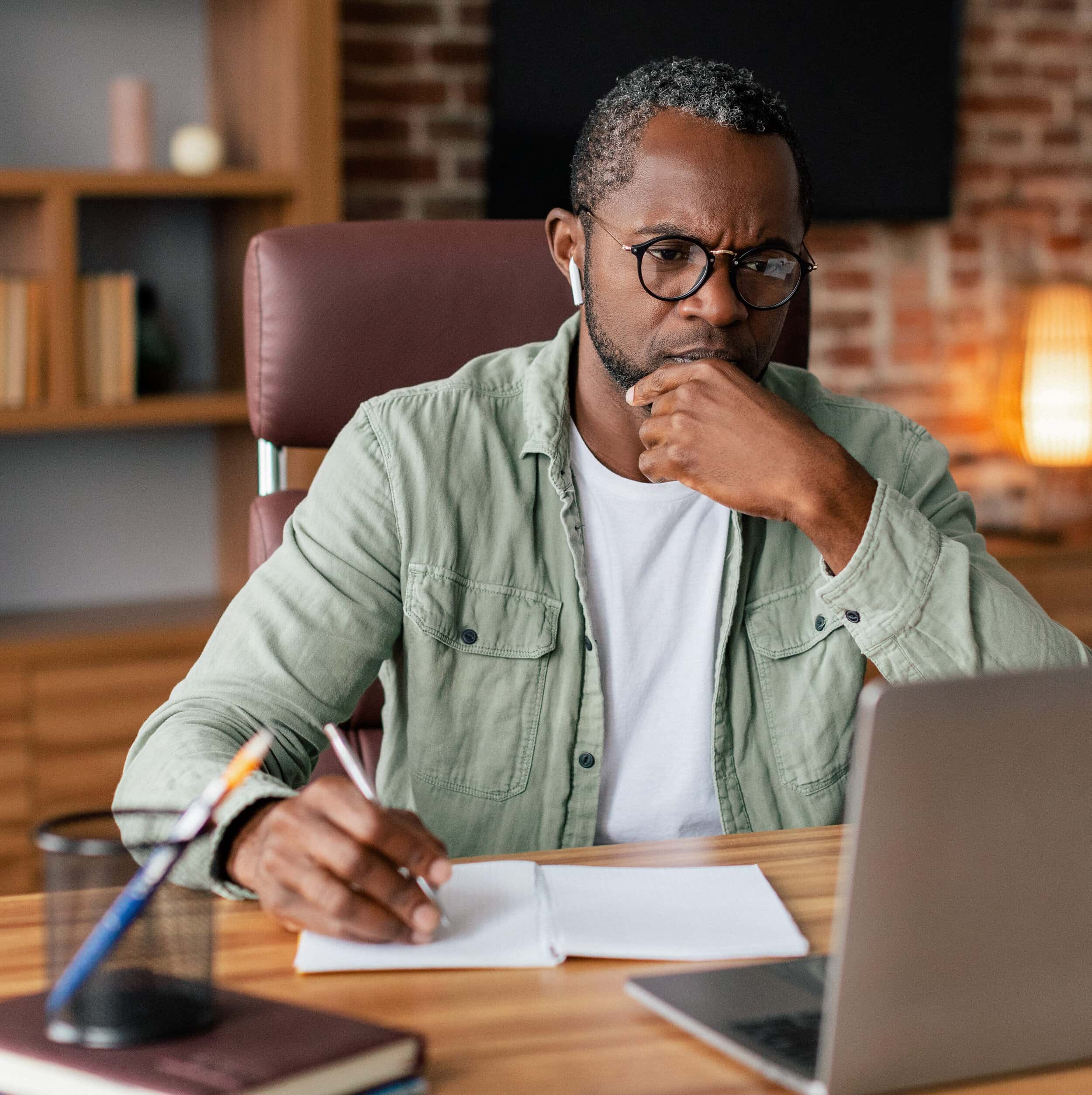 Serious concentrated adult african american man in glasses and casual in wireless headphones works on laptop