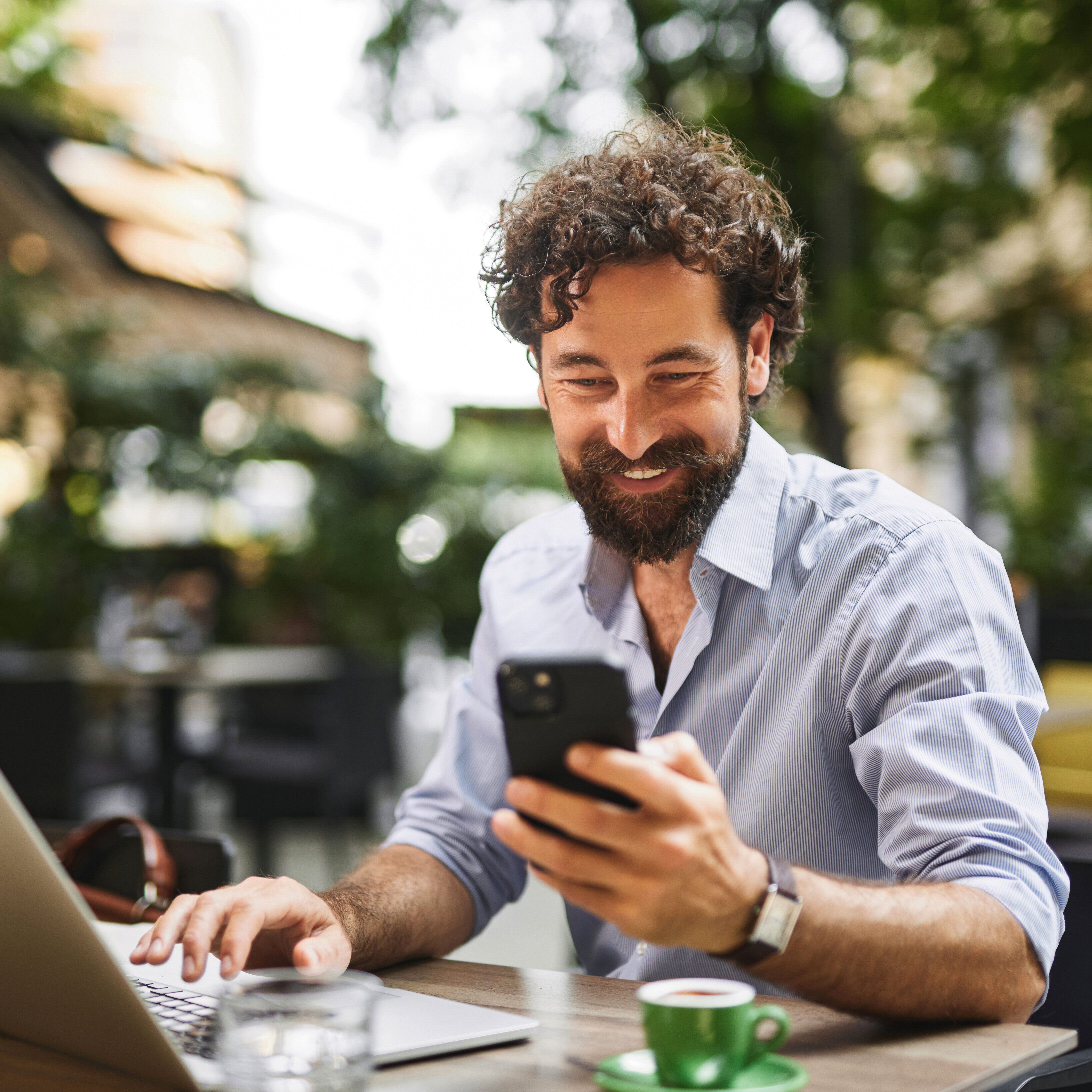 Professional working outdoors on a laptop, engaging with smartphone, enjoying coffee at a lively cafe during daytime