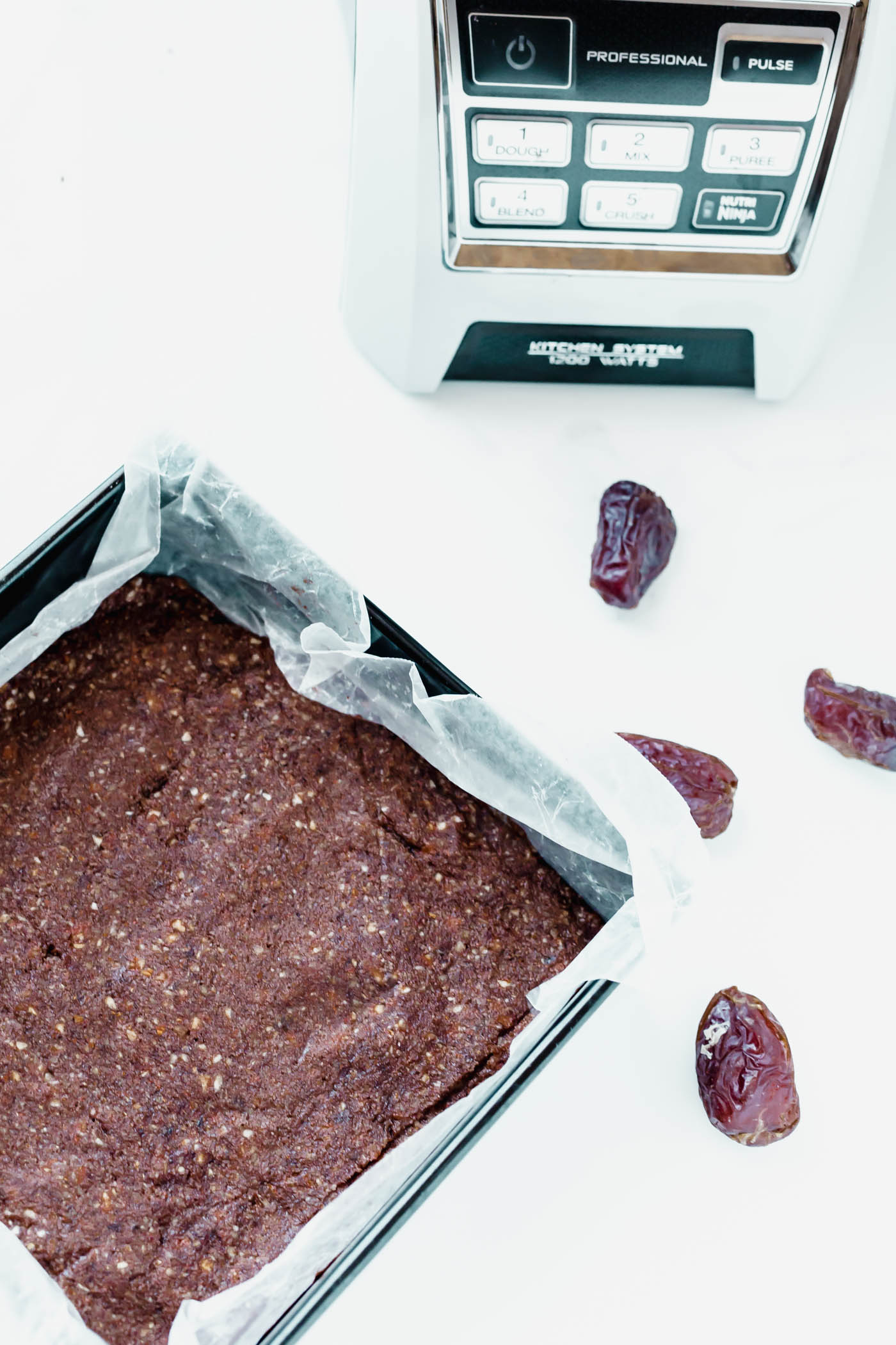 Chocolate larabars in a baking pan with dates and food processor in the background
