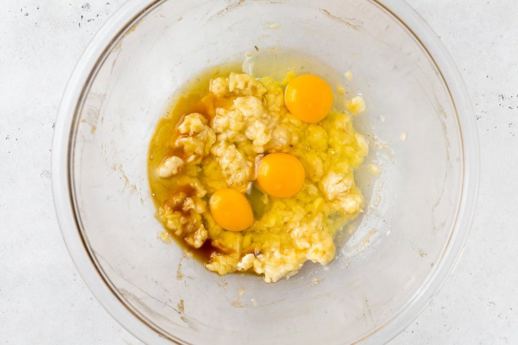 smashed banana, whole eggs, honey, and vanilla in glass mixing bowl