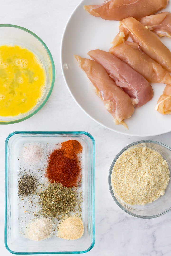 Whisked egg in bowl, raw chicken tenders, seasonings, and almond flour in bowl