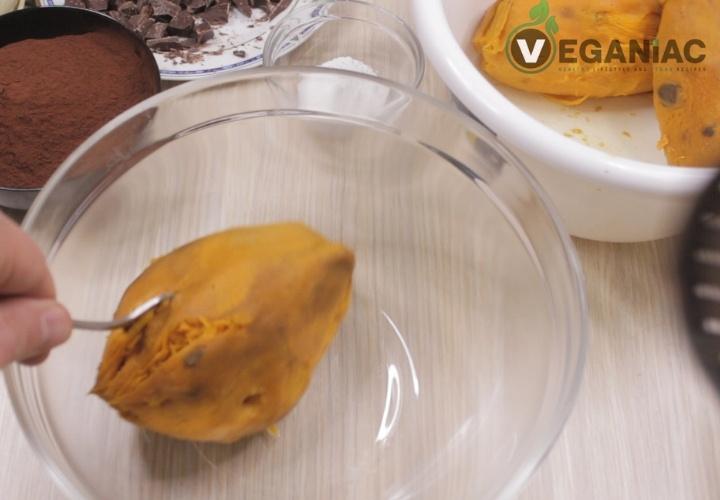 A hand holds a spoon, scooping sweet potato into a clear mixing bowl, preparing for a Sweet Potato Chocolate Cake. Ingredients like chocolate, cocoa, and sugar are scattered around. Nearby, a bowl with the "Veganac" logo peeks into view.