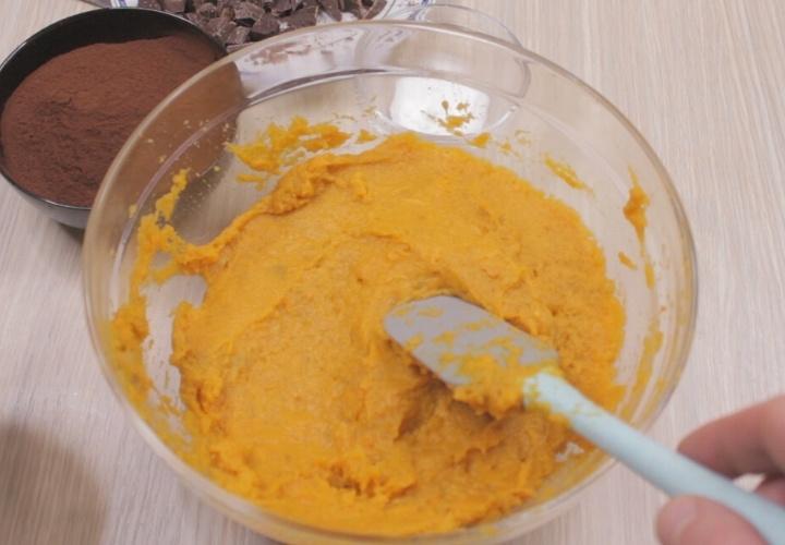A bowl filled with orange pumpkin puree is being mixed with a spatula, hinting at the start of a delicious sweet potato chocolate cake. Nearby, bowls brim with rich cocoa powder and chocolate pieces on a light wooden surface, ready to elevate your dessert to new heights.