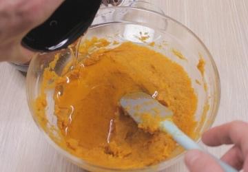 A person pours liquid from a black container into a clear bowl filled with an orange sweet potato mixture. A blue-handled spatula rests in the bowl, ready to craft the perfect cake. The bowl is placed on a light wooden surface, hinting at the promise of a delicious chocolate creation.