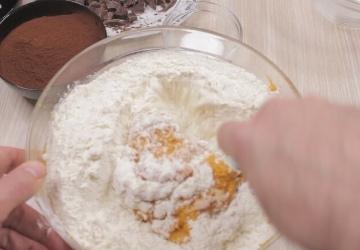 A hand mixing flour and other ingredients in a transparent bowl, with hints of brown powder and chocolate pieces for a Sweet Potato Chocolate Cake visible in the background on the light-colored countertop.