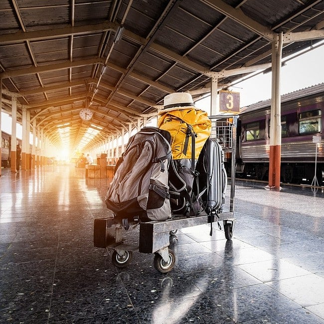 Um carrinho com duas mochilas, uma amarela e outra cinza, e um chapéu em cima, fica na plataforma de uma estação de trem com um trem visível à direita e o sol brilhando ao fundo - perfeito para quem está se perguntando como arrumar a mala para viajar.