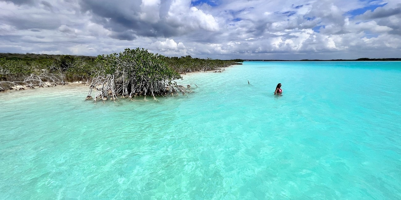 Águas cristalinas azul-turquesa com um pequeno mangue à esquerda e uma pessoa nadando à direita. O céu acima está parcialmente nublado, lembrando a beleza serena encontrada em Bacalar, no México.