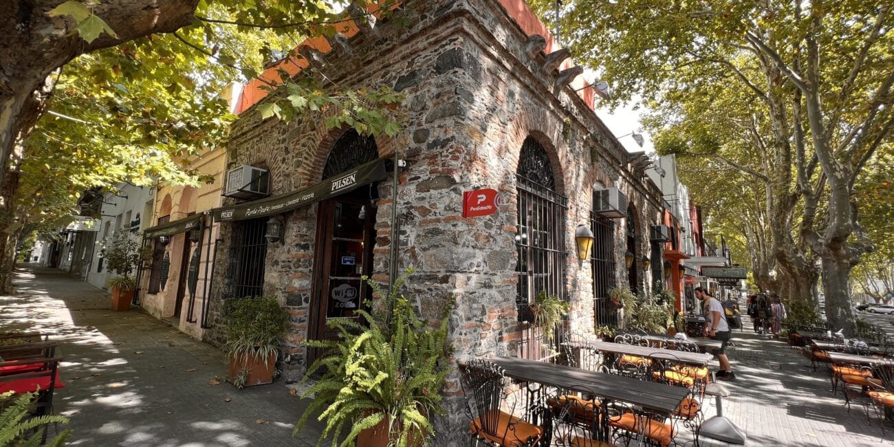 Um edifício de pedra com áreas de estar ao ar livre é sombreado por grandes árvores em Colônia del Sacramento. As mesas e cadeiras estão dispostas ao longo da calçada. Uma placa vermelha com texto branco está pendurada no prédio enquanto as pessoas caminham nas proximidades.