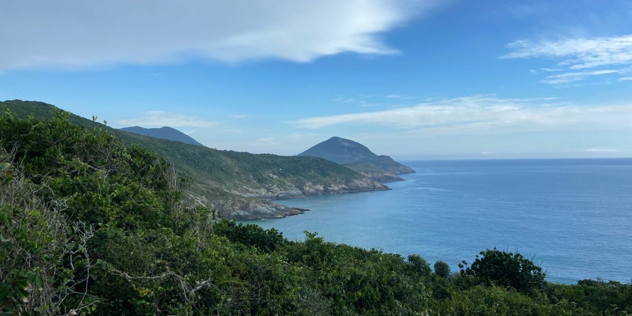 A vegetação exuberante em primeiro plano, o oceano azul e as montanhas ao fundo sob um céu parcialmente nublado criam uma vista pitoresca de Arraial do Cabo.