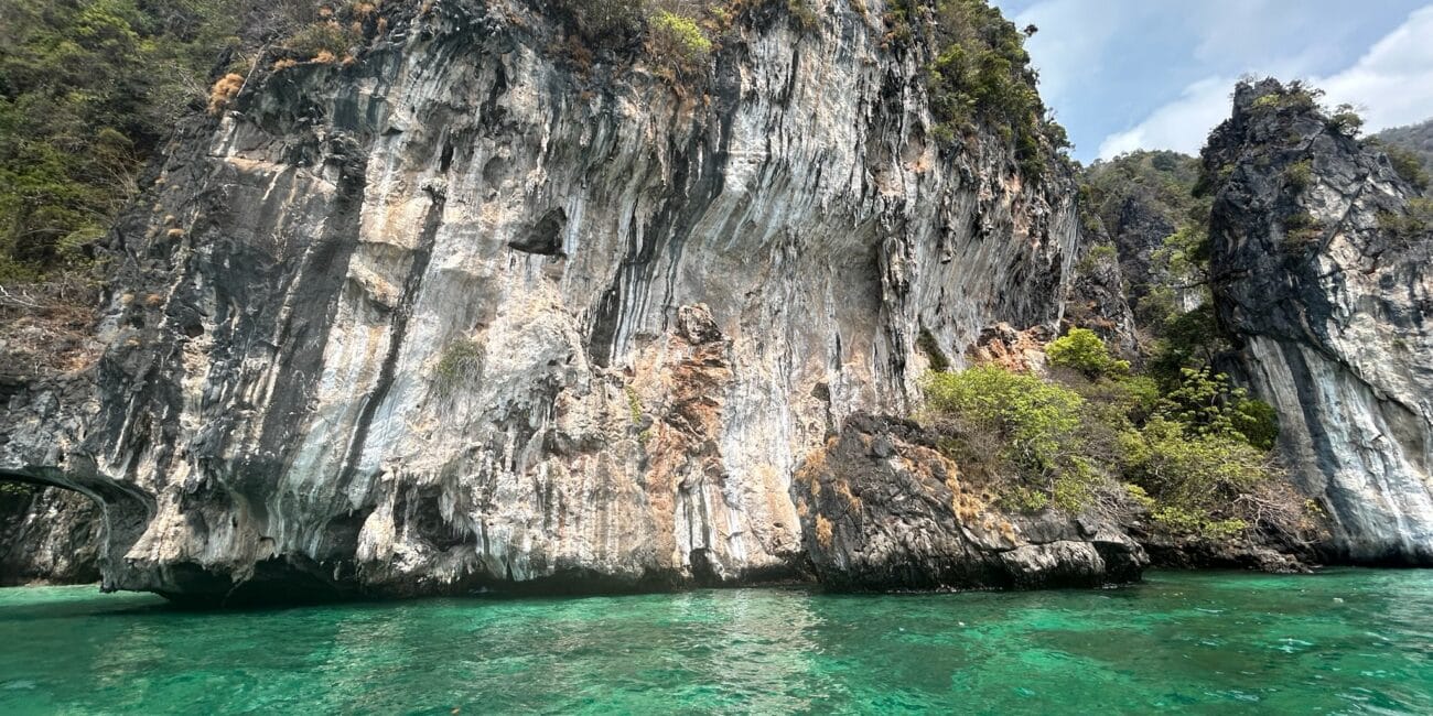 Um penhasco alto com superfícies rochosas escarpadas ergue-se ao lado de águas cristalinas azul-turquesa sob um céu parcialmente nublado. A vegetação esparsa é vista na falésia, lembrando a beleza natural frequentemente encontrada ao explorar os meios de transporte na Tailândia.