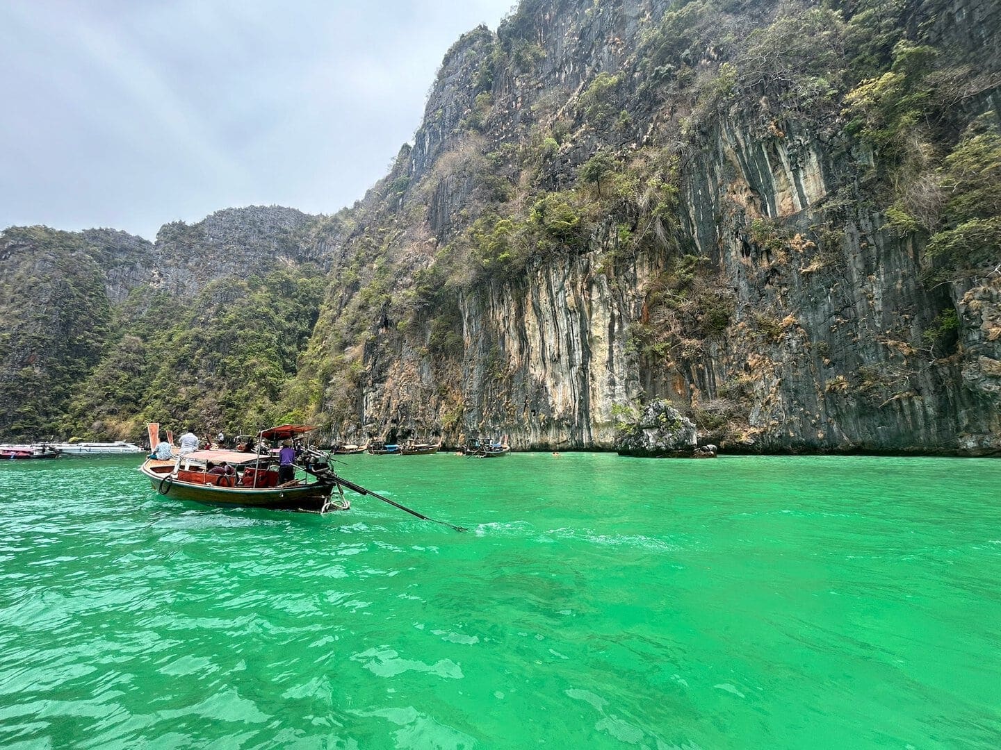 Um pequeno barco com motor de popa, meio de transporte comum em Tailândia, flutua em águas verdes perto de grandes falésias rochosas cobertas de vegetação em um dia claro.