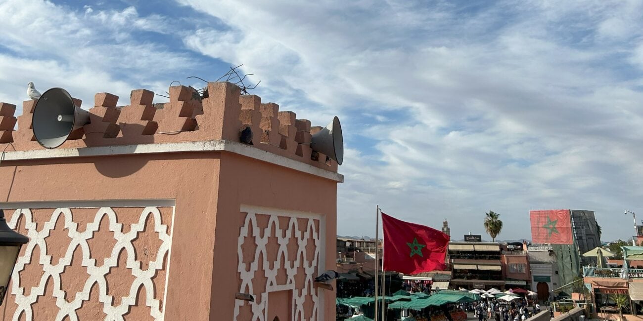 Uma vista do topo de um mercado em Marrakech no Marrocos com barracas abaixo, exibindo uma grande bandeira vermelha com uma estrela verde contra um céu azul pontilhado de nuvens. Um edifício com padrão geométrico rosado se destaca em primeiro plano.