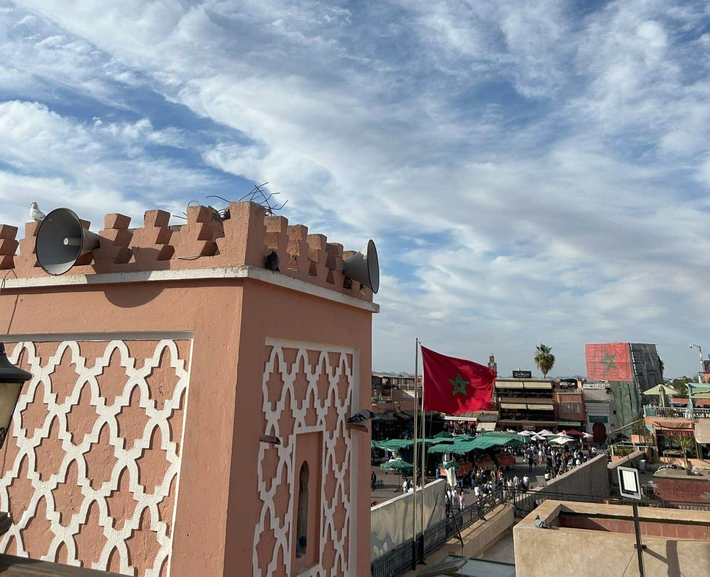 Uma vista do topo de um mercado em Marrakech no Marrocos com barracas abaixo, exibindo uma grande bandeira vermelha com uma estrela verde contra um céu azul pontilhado de nuvens. Um edifício com padrão geométrico rosado se destaca em primeiro plano.