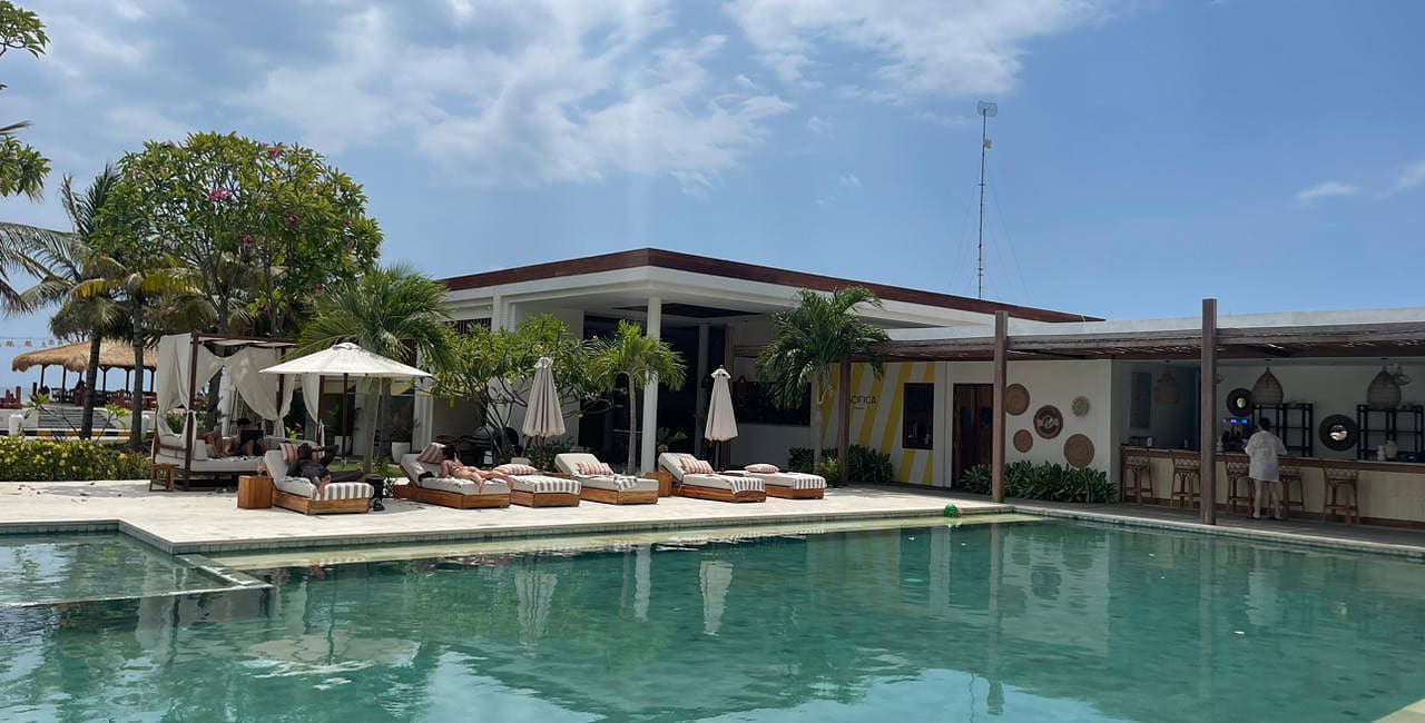 Área de piscina externa com espreguiçadeiras, guarda-sóis e palmeiras ao lado de um edifício moderno sob um céu parcialmente nublado - um local ideal para quem procura onde ficar em Lombok na Indonésia: melhores hotéis.