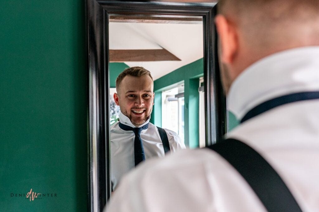 Groom adjusting his tie and looking at himself in a mirror before the wedding at Wasing Park, captured by Surrey wedding photographer Denise Winter.
